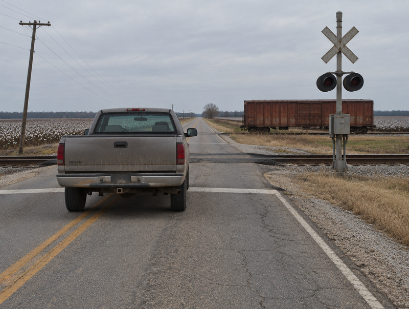 A dusty pickup truck waits at a rural level crossing with a crossbuck, signal mast, and a boxcar on a siding.