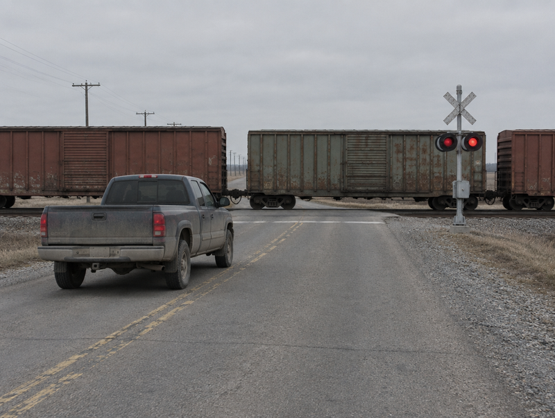 A dusty pickup truck with its windows closed waits at a rural level crossing as freight boxcars roll past.