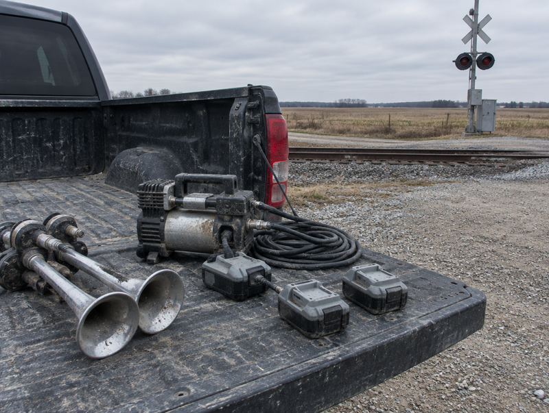 A dusty pickup tailgate by a rail crossing holds train horns, a small air compressor and three cordless tool batteries.