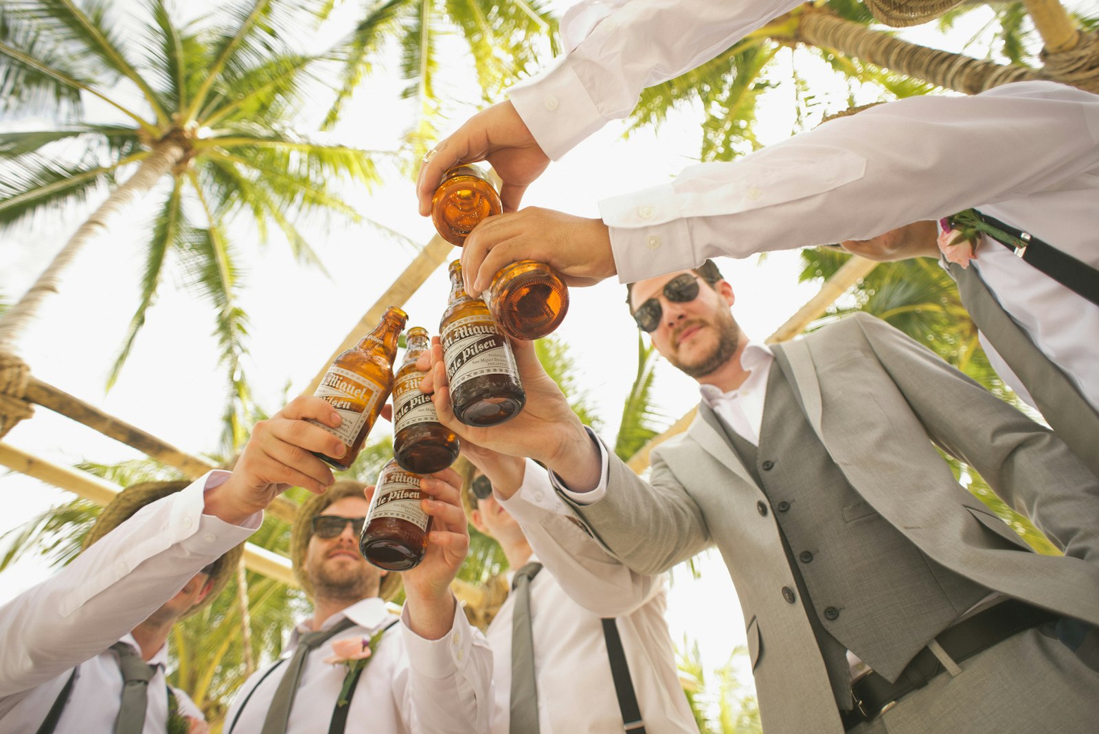 Low-angle view of a group of men in dress shirts and vests clinking bottles in a toast outdoors under palm trees, celebrating together