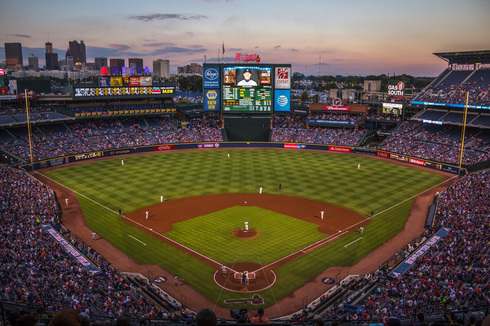 A sunny baseball diamond seen from the stands with players on the infield grass and a crowd of fans filling the seats on a clear afternoon