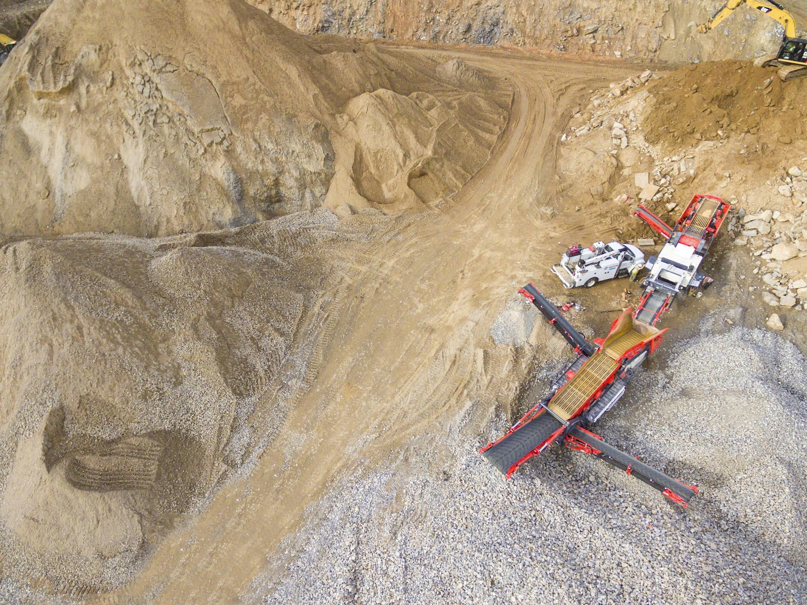 Overhead view of an open-pit quarry with heavy excavation equipment and track marks across layered tan and grey rock