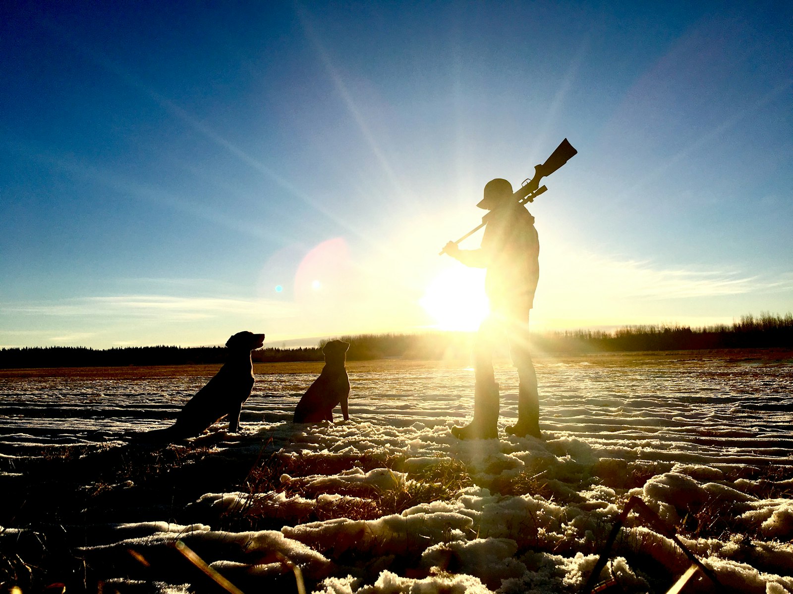 A hunter in the field standing with a rifle and his hunting dogs on open snowy terrain, the kind of outdoor setting where a loud signal horn helps partners stay in contact