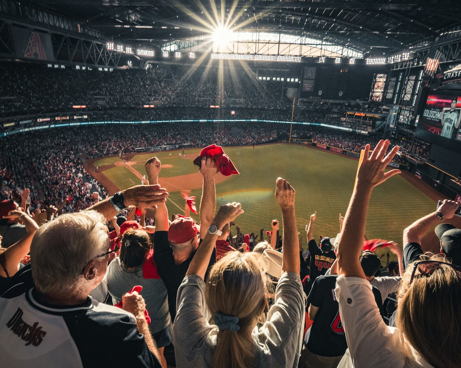 A crowd of fans packed into the stadium stands, standing with arms raised and cheering together on a sunny game day