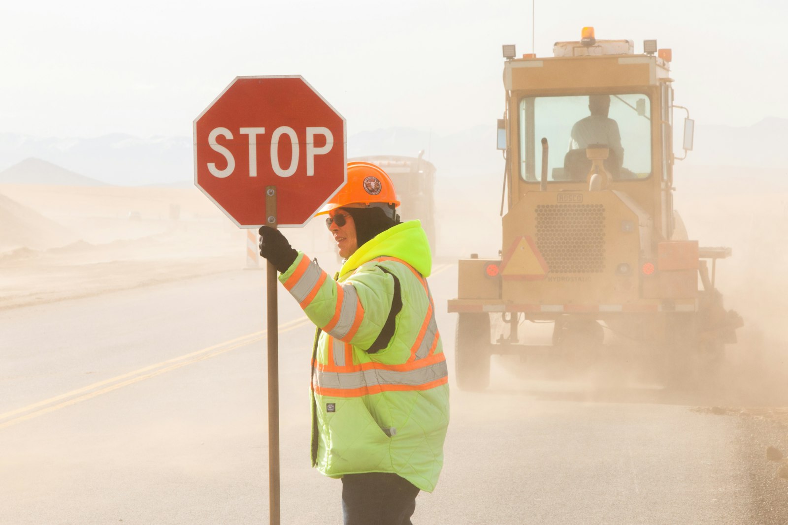 A flagger in a high-visibility green and yellow safety jacket and hard hat holding a red stop sign on a highway road-work project, with heavy equipment in the background