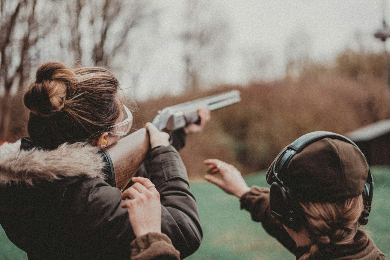 A shooter at an outdoor range aiming a rifle at a downrange target while wearing ear protection, with an instructor standing alongside