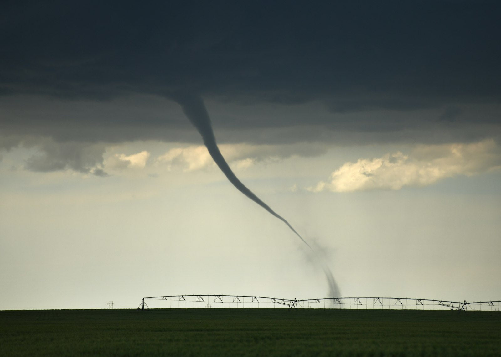 A funnel cloud touching down near an irrigation pivot in an open farm field under heavy grey storm clouds