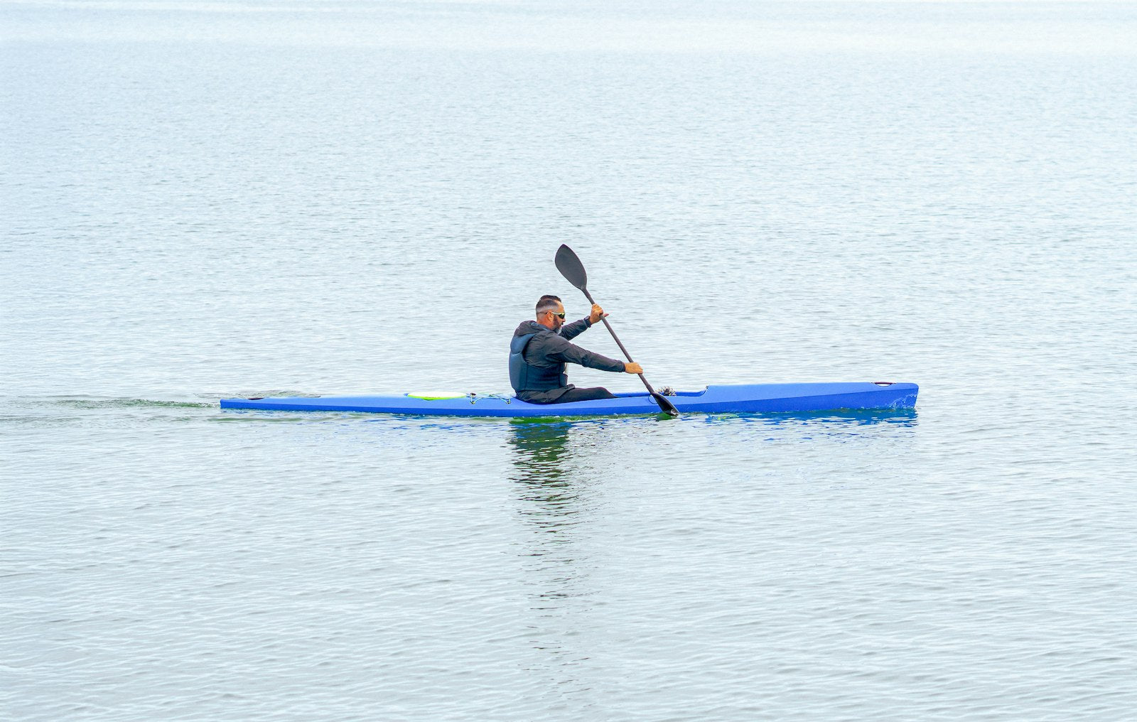 A paddler propelling a blue kayak across calm open water, paddle mid-stroke, on a bright clear day