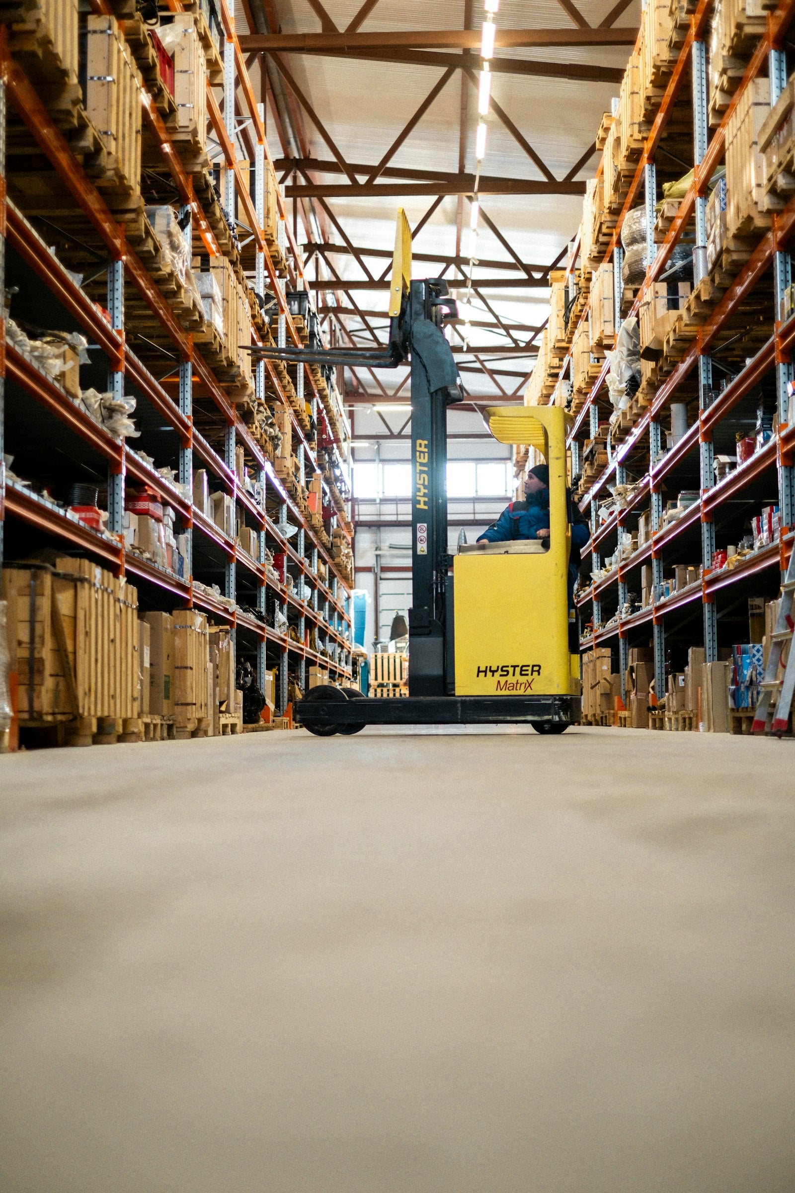 A yellow forklift parked in a large warehouse aisle lined with tall steel pallet racking stacked with boxes