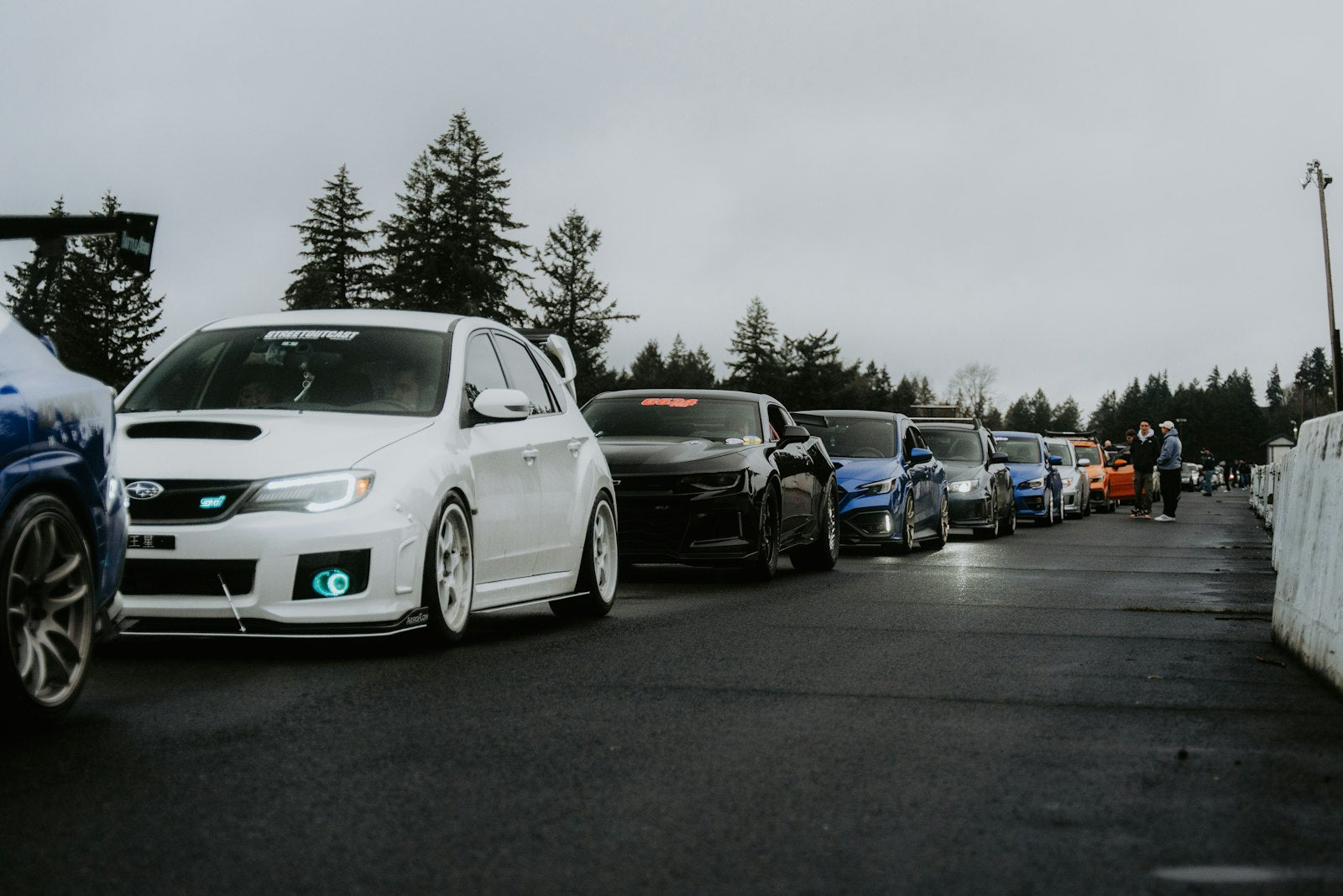 A long row of modified cars lined up wheel-to-wheel at an outdoor car meet, hoods and rims on display under open sky