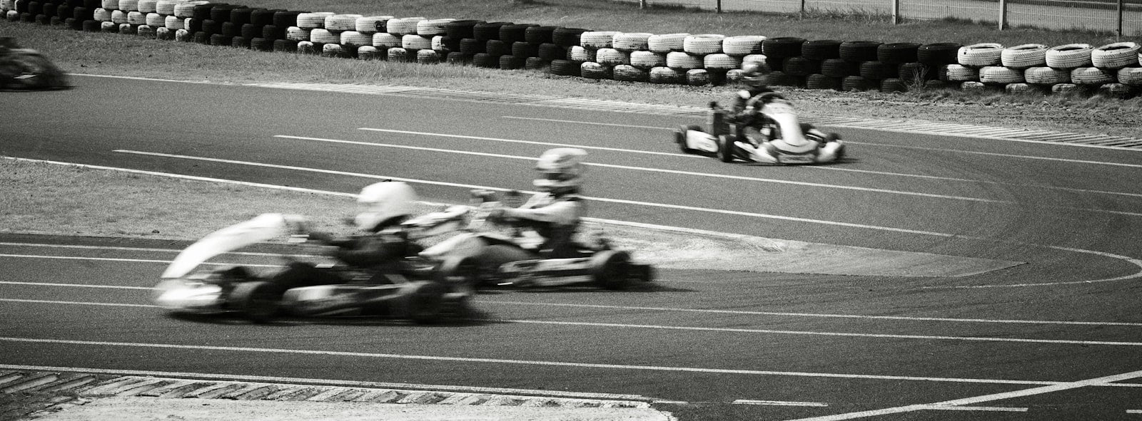 A pack of go-karts racing wheel-to-wheel down an outdoor track lined with safety barriers