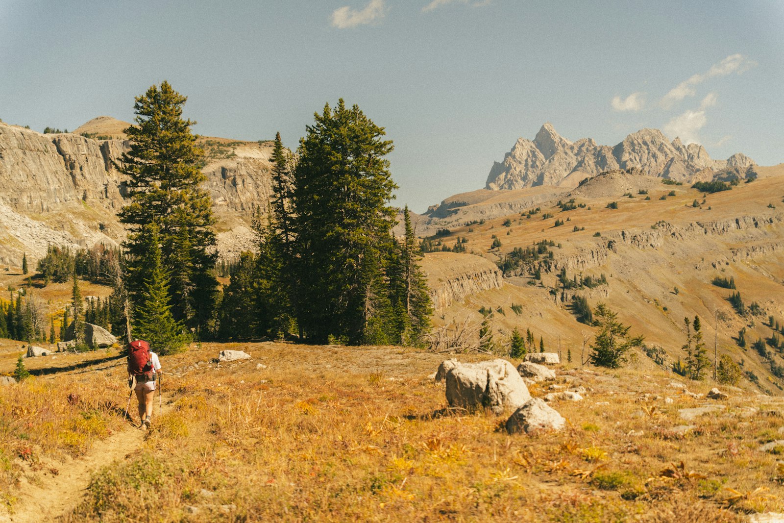 A hiker with a loaded backpack walking a rocky mountain trail toward distant forested peaks under open sky