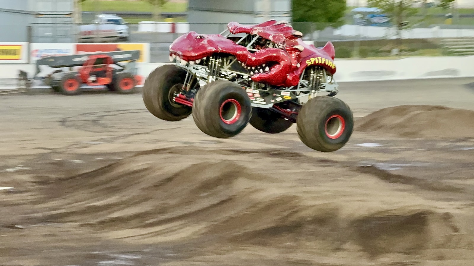 Bright red monster truck airborne mid-jump over a dirt mound at an outdoor motorsport event