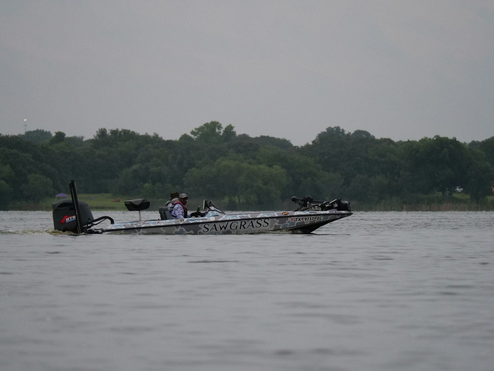 A bass angler in a fishing boat moving across a calm early-morning lake, the kind of open water where a tournament takeoff horn carries