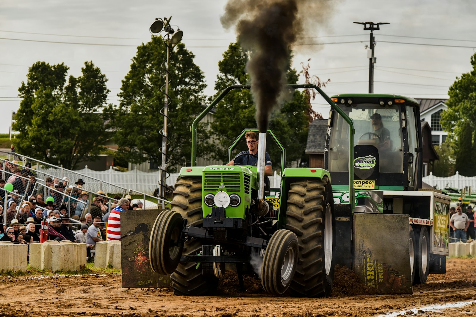 A powerful green pulling tractor dragging a weighted sled down a dirt track, throwing black exhaust smoke at an outdoor truck and tractor pull